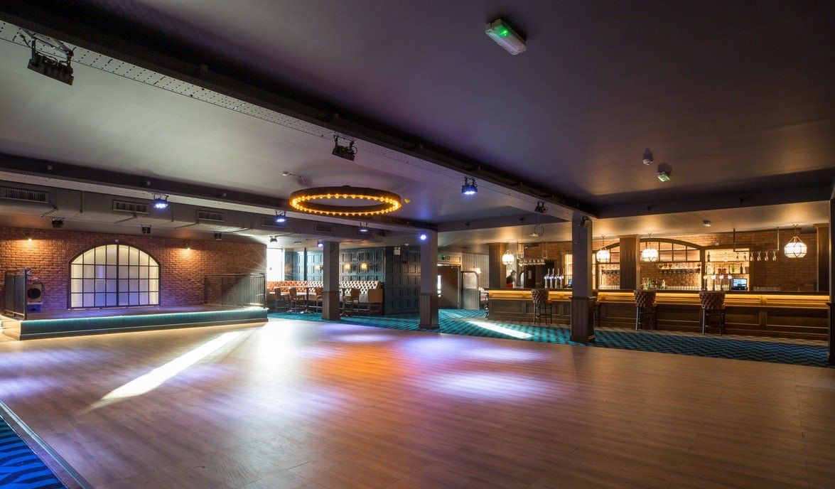Empty ballroom with wood floor, chandeliers, arched windows, and a lit bar along the right wall.