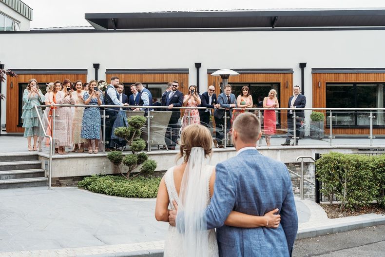 Bride and groom watching a wedding party gathered on a patio outside a modern building