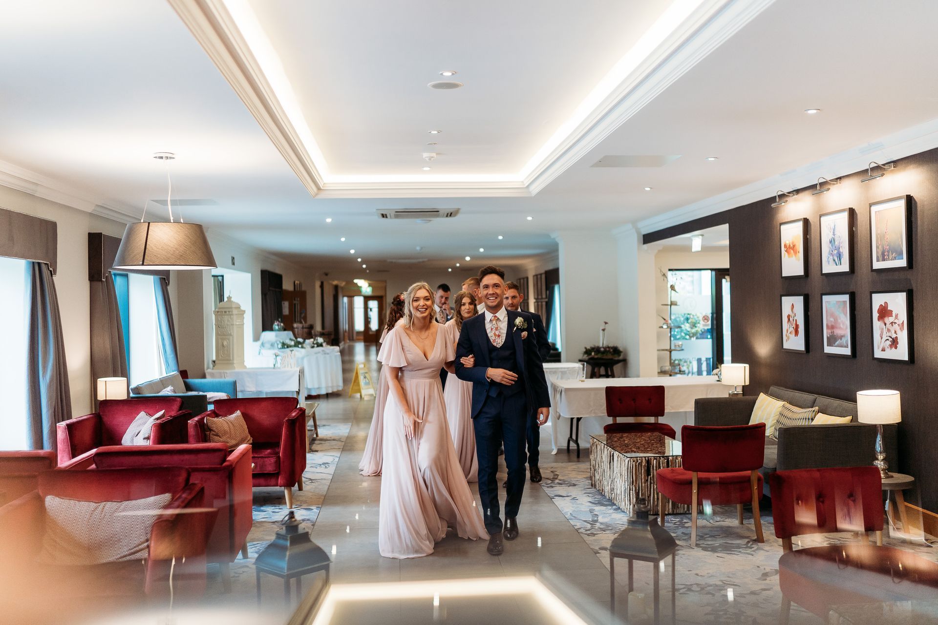 Bride and groom walking through a bright hotel lobby with red seating and framed wall art