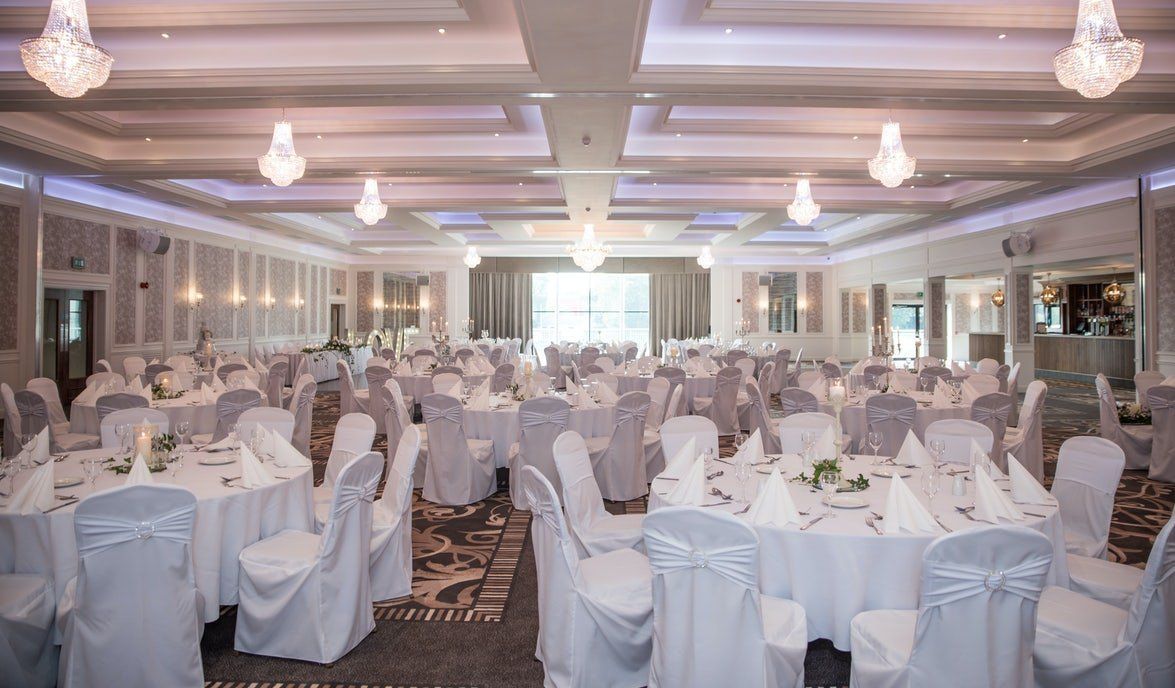 Elegant banquet hall set with round tables, white chair covers, and chandeliers under soft lighting.
