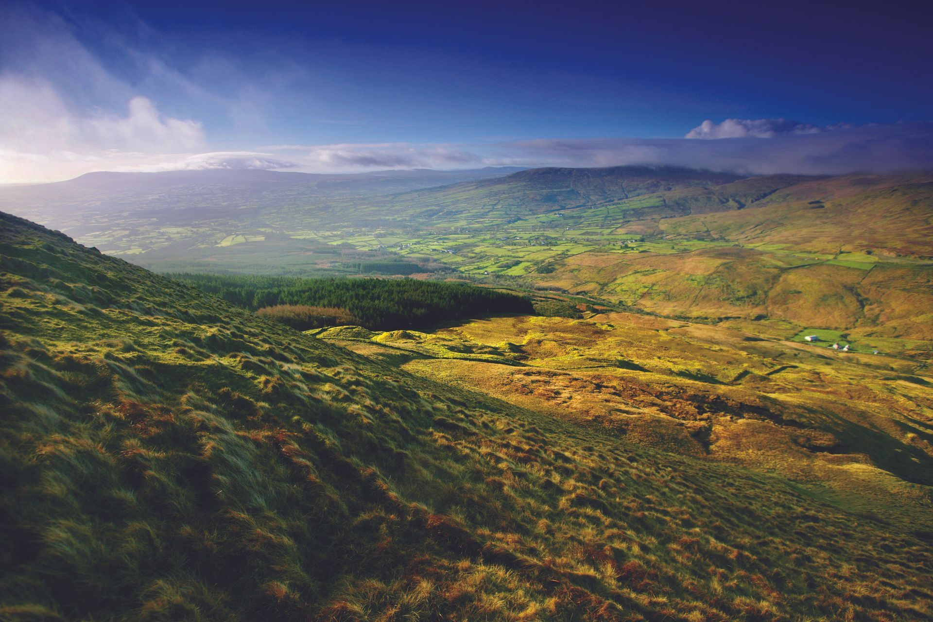 Wide mountain valley with green hills, golden fields, and a cloudy sky at sunrise