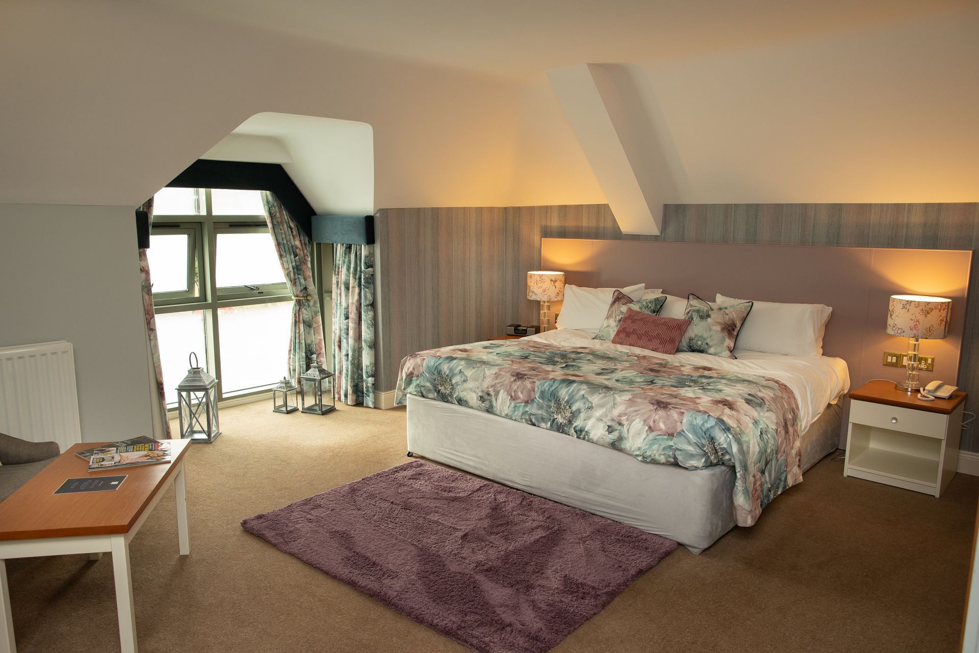 Spacious bedroom with a large bed, purple rug, desk, and window seating in warm neutral tones