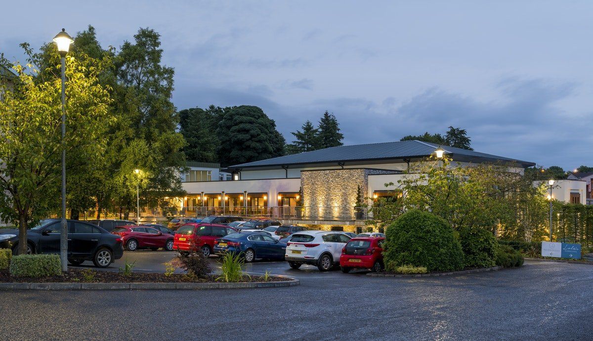 Parking lot outside a lit building at dusk, with cars and trees in front.