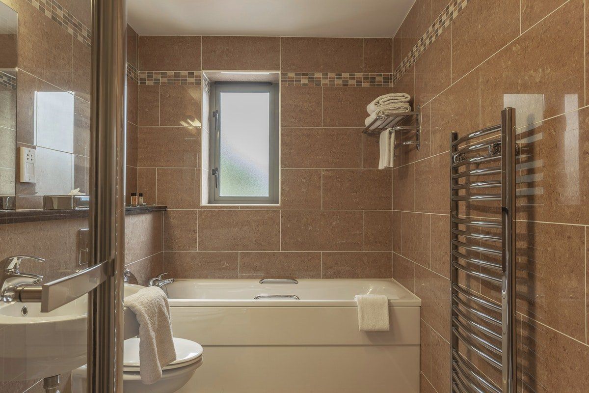 Brown-tiled bathroom with bathtub, sink, mirror, window, and heated towel rail.