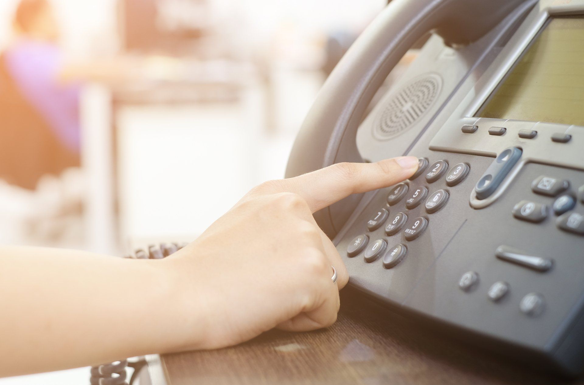 Finger pressing buttons on a gray office phone handset on a desk