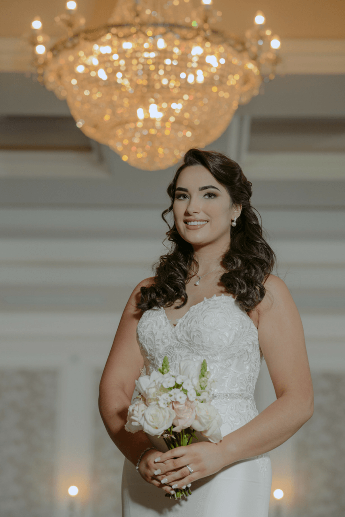 Bride in a white dress holding a bouquet, standing indoors beneath a chandelier and smiling.