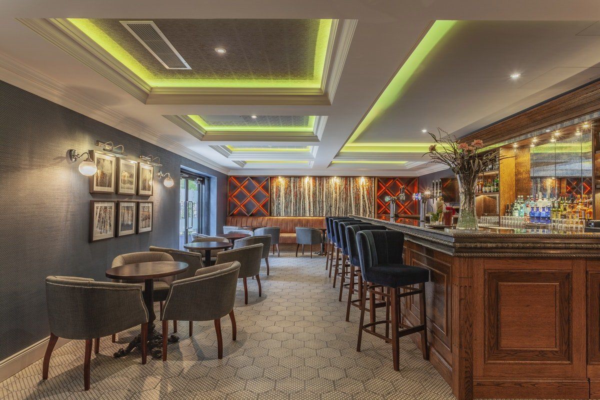 Empty upscale bar and lounge with wooden counter, high stools, and green-lit ceiling panels