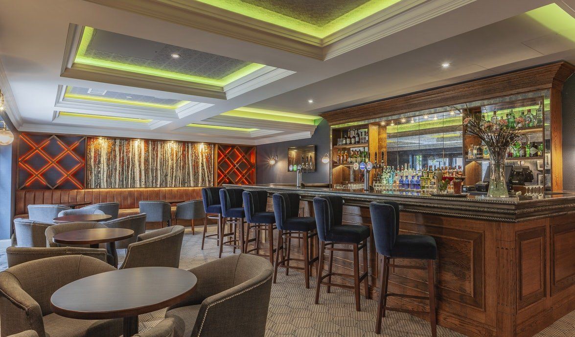 Modern hotel bar and lounge with stools, round tables, wood counter, and neon-lit ceiling panels