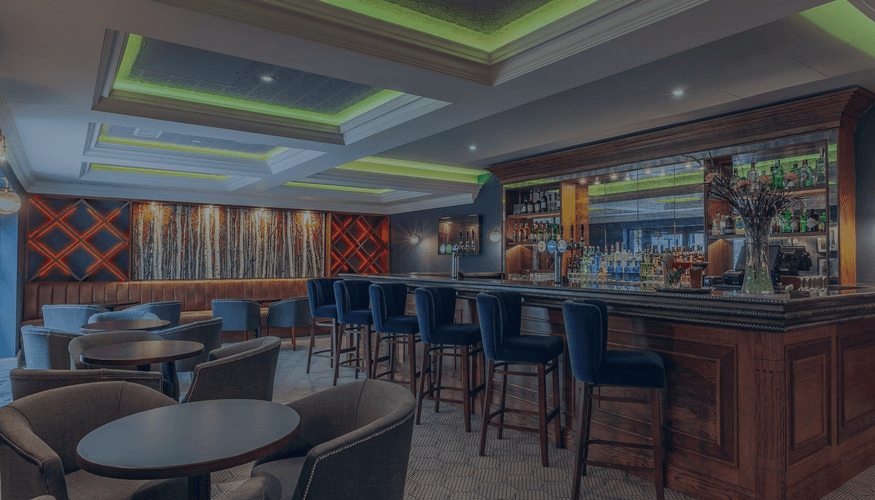 Modern bar lounge with blue stools, wooden counter, tables, and neon-accented ceiling lighting