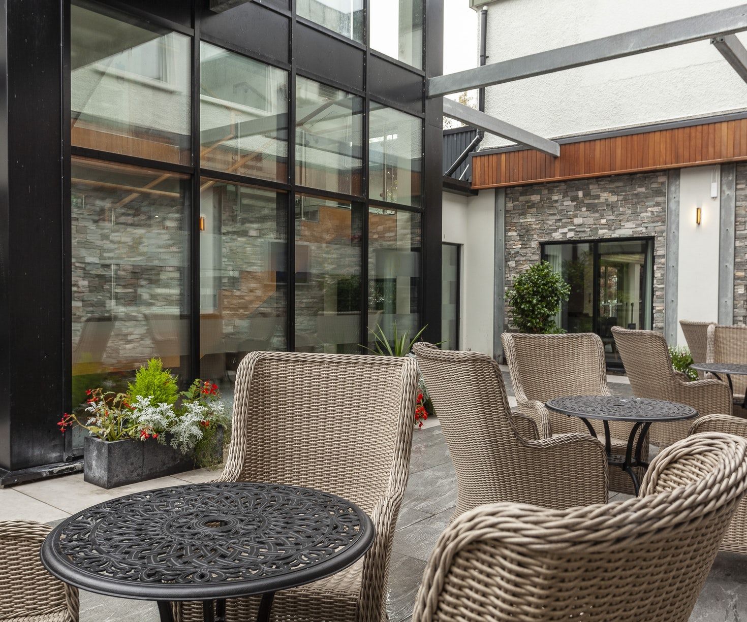 Modern outdoor café patio with wicker chairs, round tables, glass walls, and potted flowers