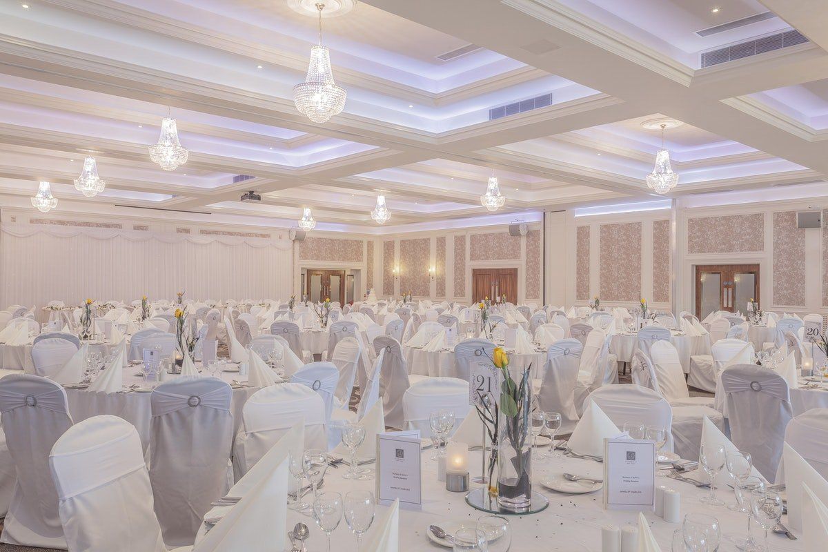 Elegant banquet hall with white table settings, draped chairs, and chandeliers under a lit ceiling