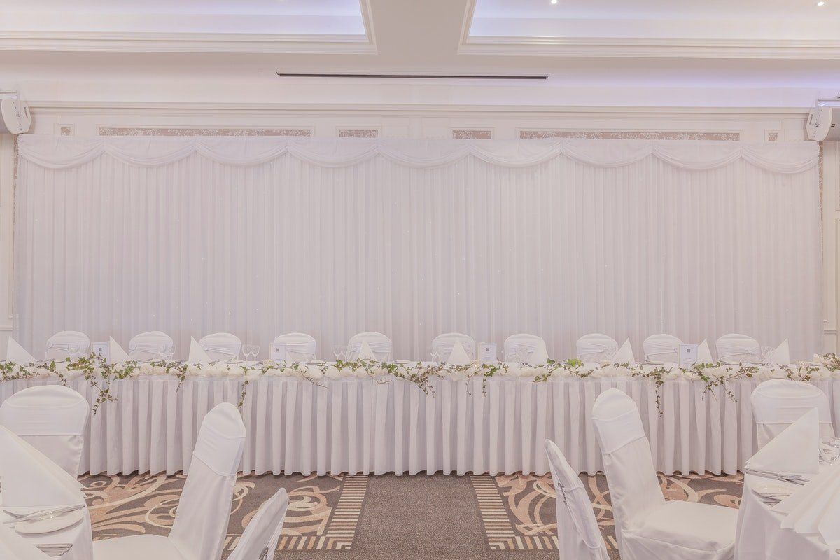 White floral wedding reception backdrop with draped curtains and decorated banquet table in a bright hall