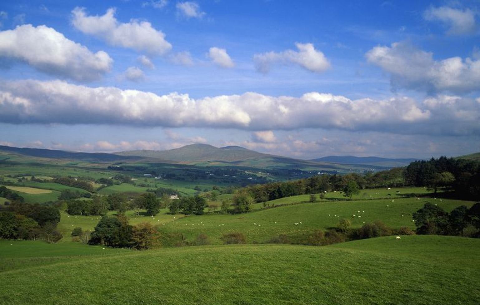 Green rolling hills under a blue sky with scattered clouds and distant mountains