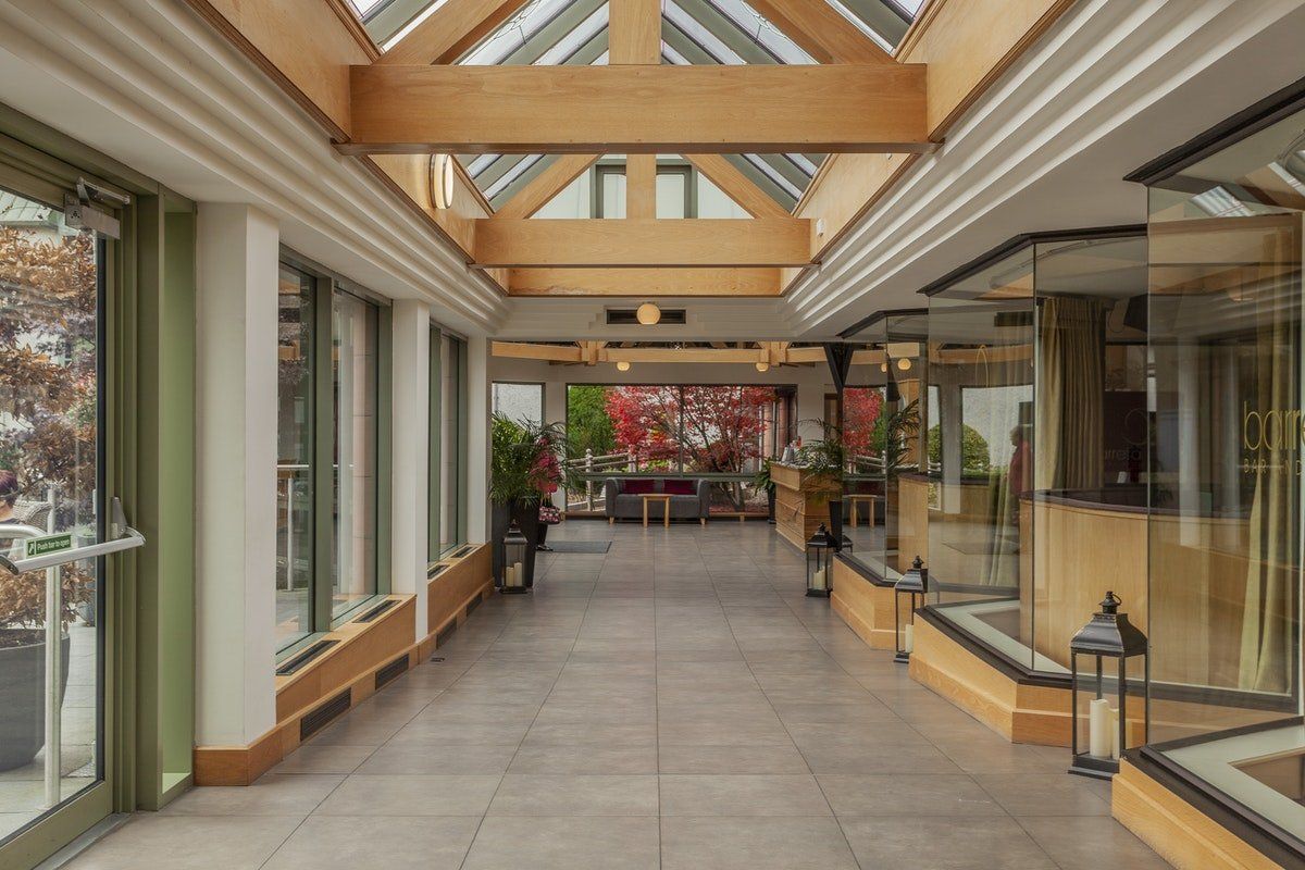 Modern indoor corridor with glass storefronts, skylights, potted plants, and tiled floor