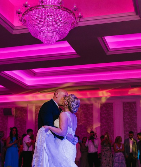 Couple dancing and embracing under pink-lit chandeliers at a formal reception
