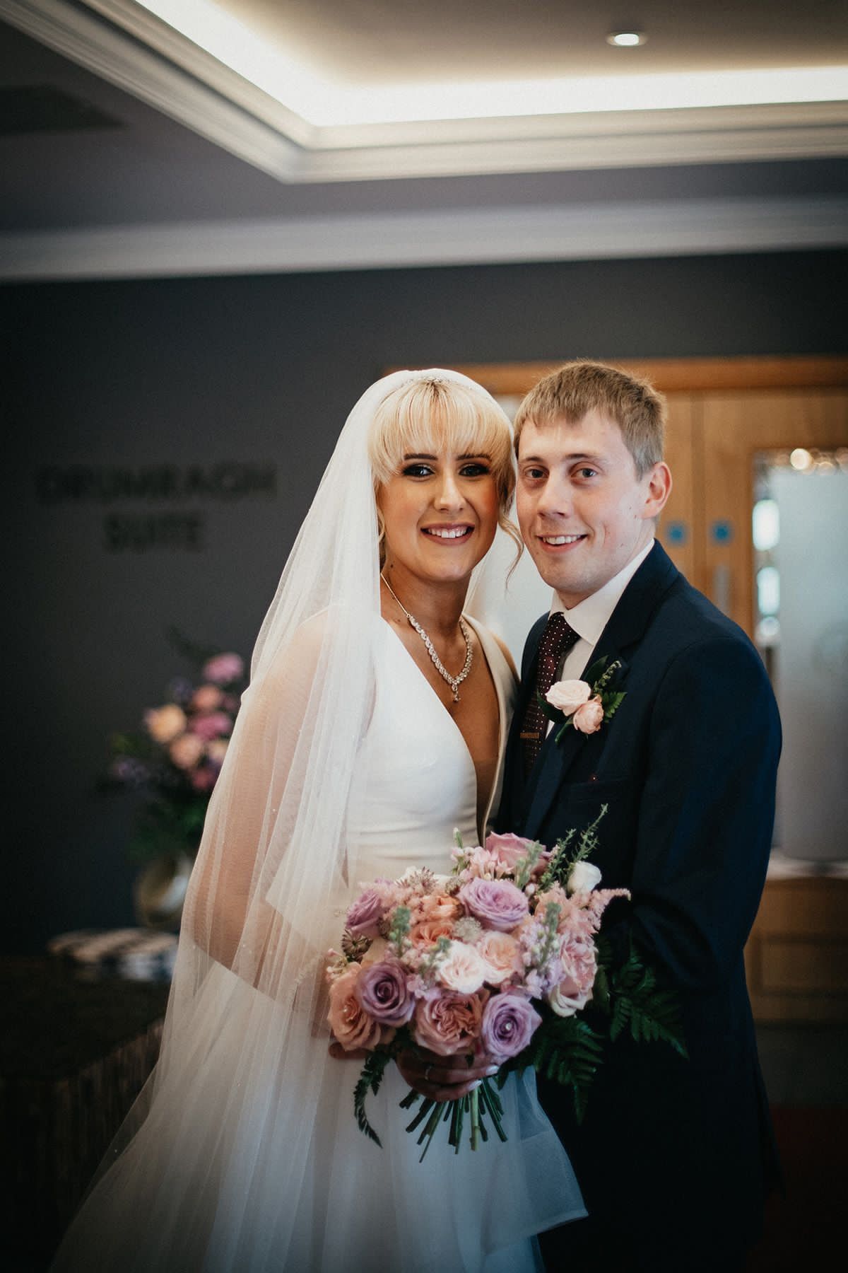 Bride and groom posing indoors, holding a bouquet, with smiling expressions and soft wedding lighting