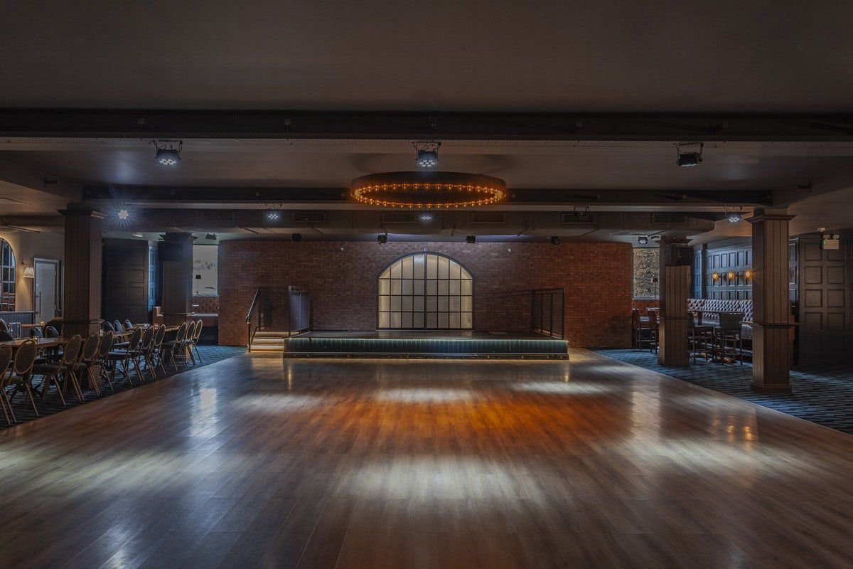 Empty modern event hall with wooden floor, brick walls, arched windows, and a chandelier overhead
