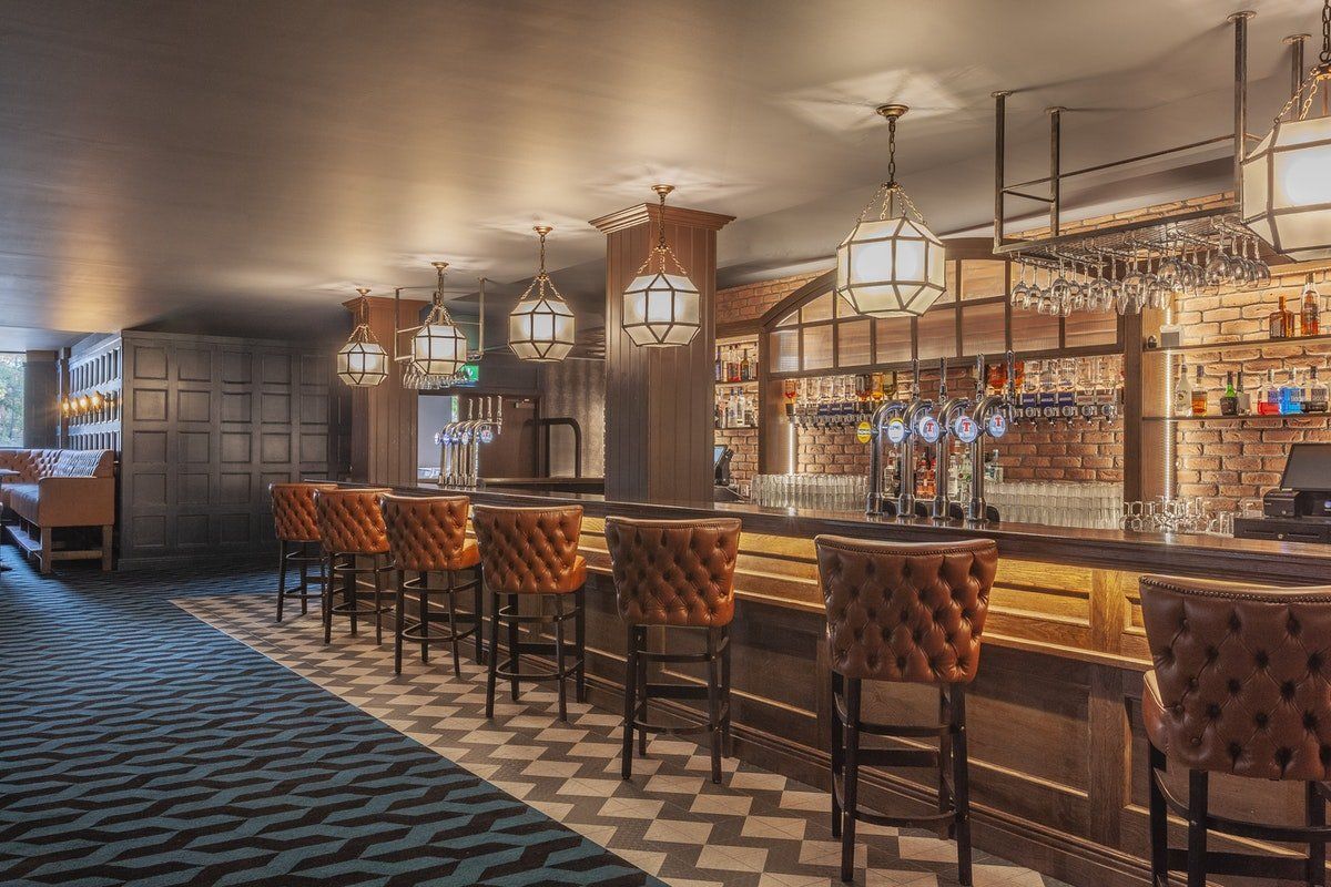Warmly lit vintage bar interior with a long wooden counter, tufted stools, and hanging pendant lights