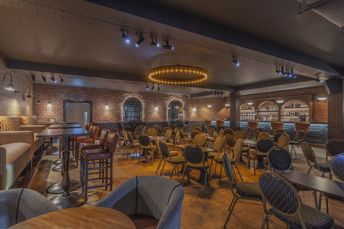 Dimly lit modern lounge with patterned chairs, tables, brick walls, and a circular ceiling light.