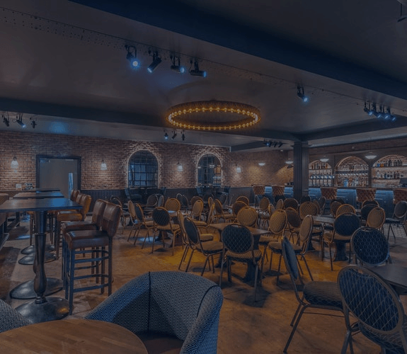Dimly lit lounge with tables and chairs, brick walls, and a glowing oval chandelier over a wooden floor.