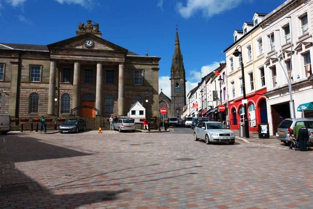 Town square with a historic stone church, clock tower, and white buildings with red storefronts on a cobblestone street