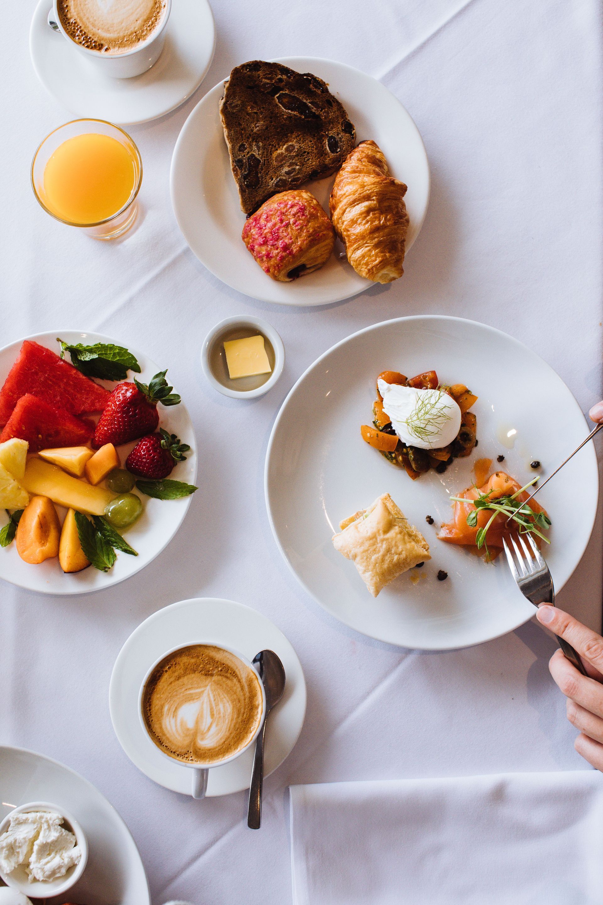 Breakfast spread with pastries, fruit, coffee, and a hand serving a bite from a plate