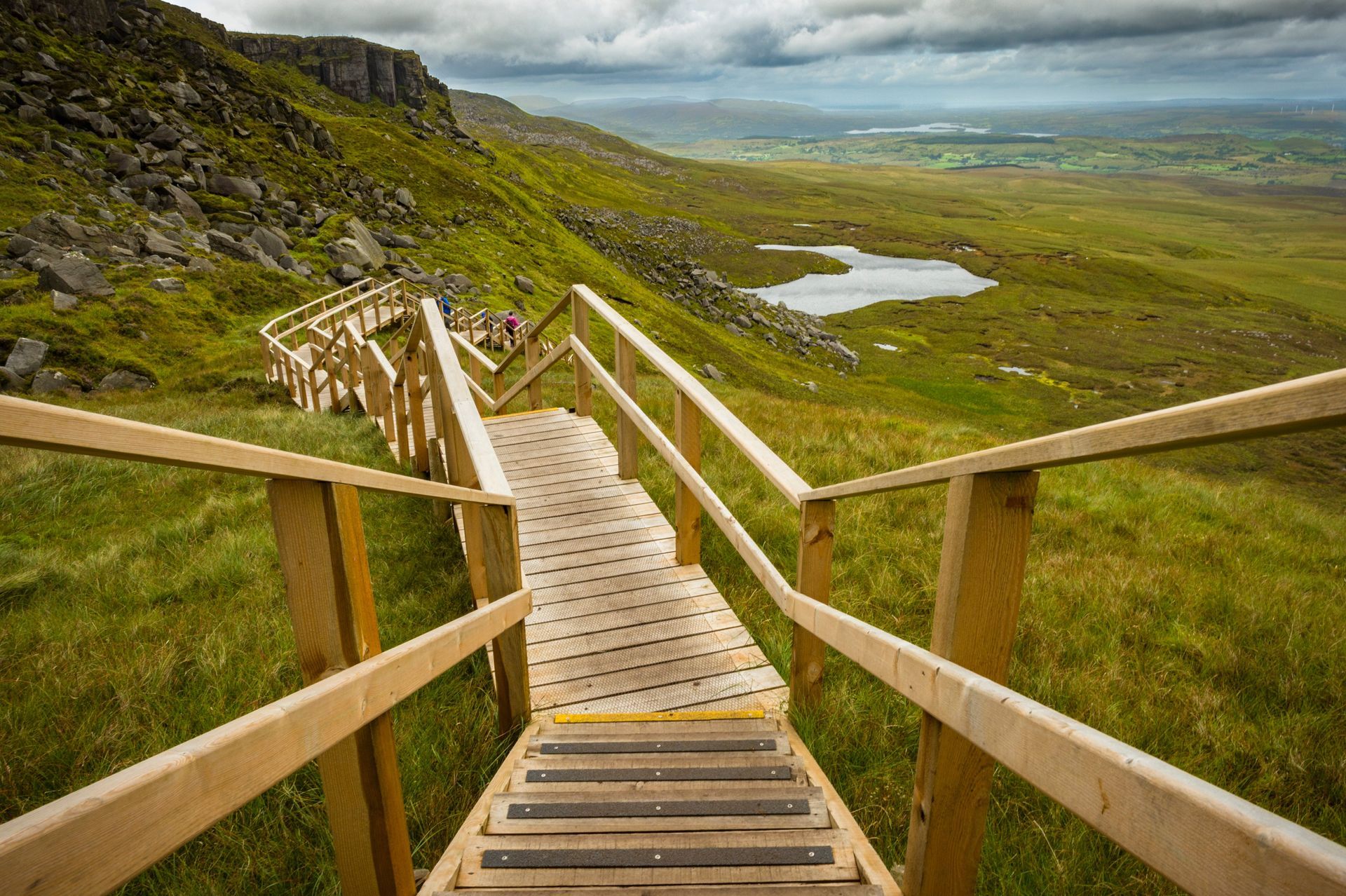 Wooden stairway descending through green hills toward a valley with a small lake under cloudy skies