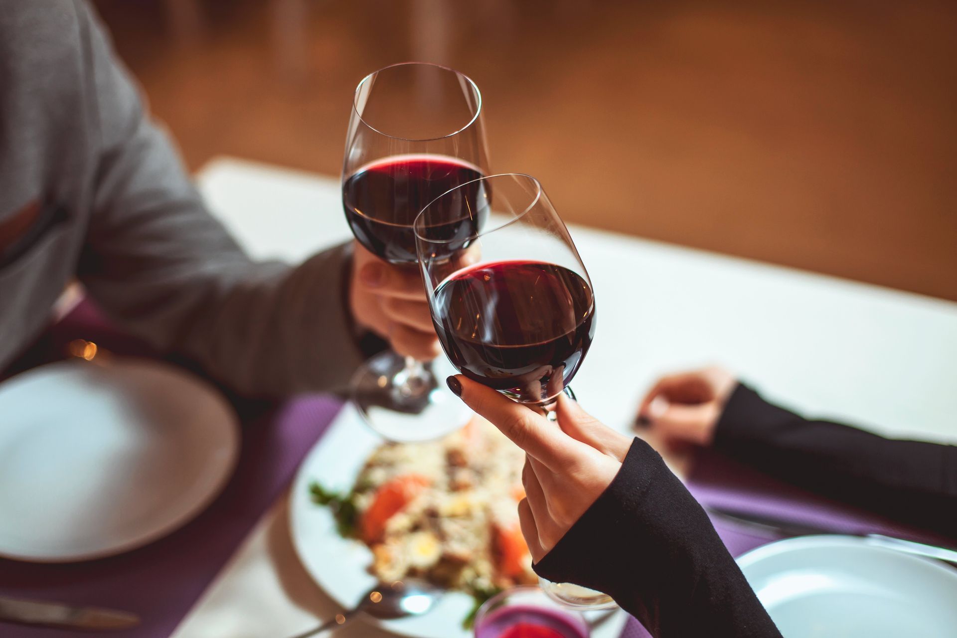 Two people toasting with red wine glasses over a restaurant dinner table
