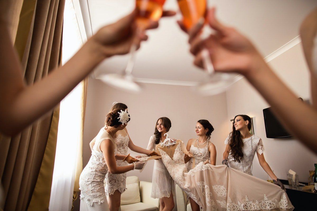 Bridesmaids in white dresses toasting champagne at a bridal getting-ready party, with a bride in the background