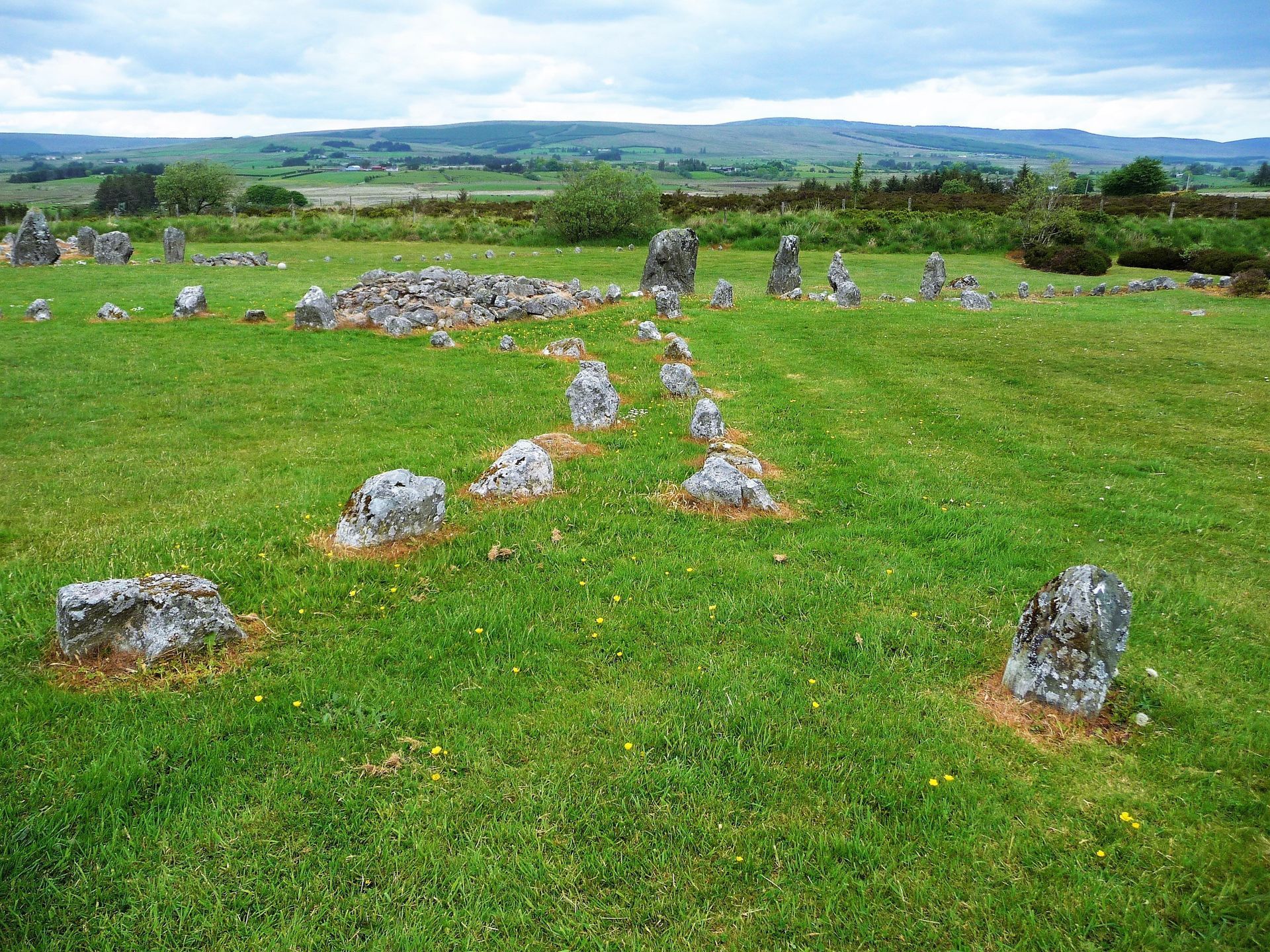 Stone circle in a green field with scattered rocks and distant hills under a cloudy sky
