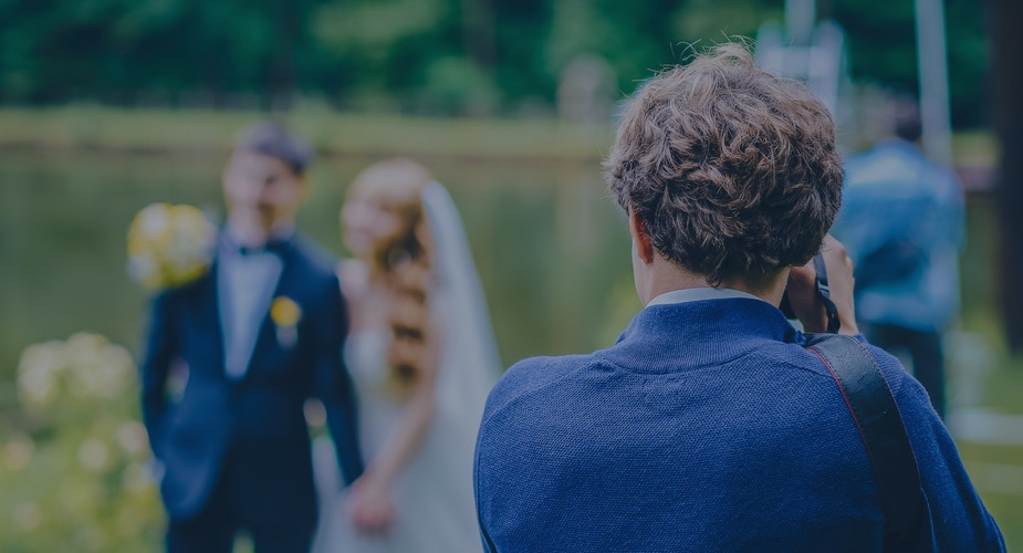 Photographer taking a wedding photo of a bride and groom outdoors, with the couple softly blurred in the background