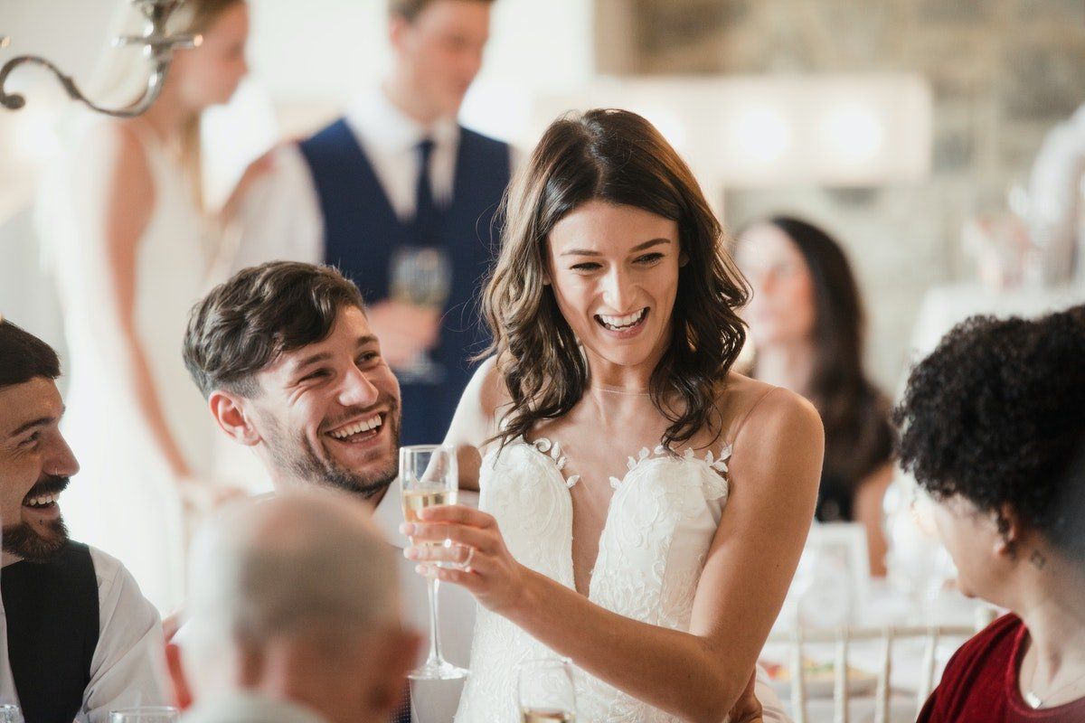 Smiling guests toasting at a wedding reception, with a woman in a white dress holding a drink.