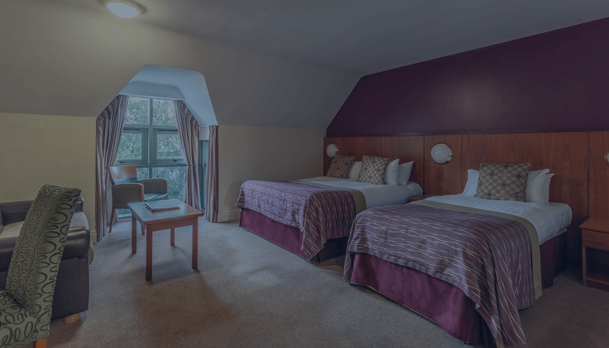 Hotel room with two beds, a chair, and a window view, lit in soft purple tones