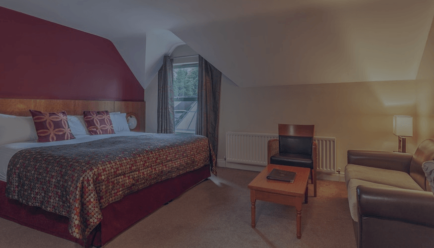 Modern hotel room with a bed, armchair, desk, lamp, and window seating area