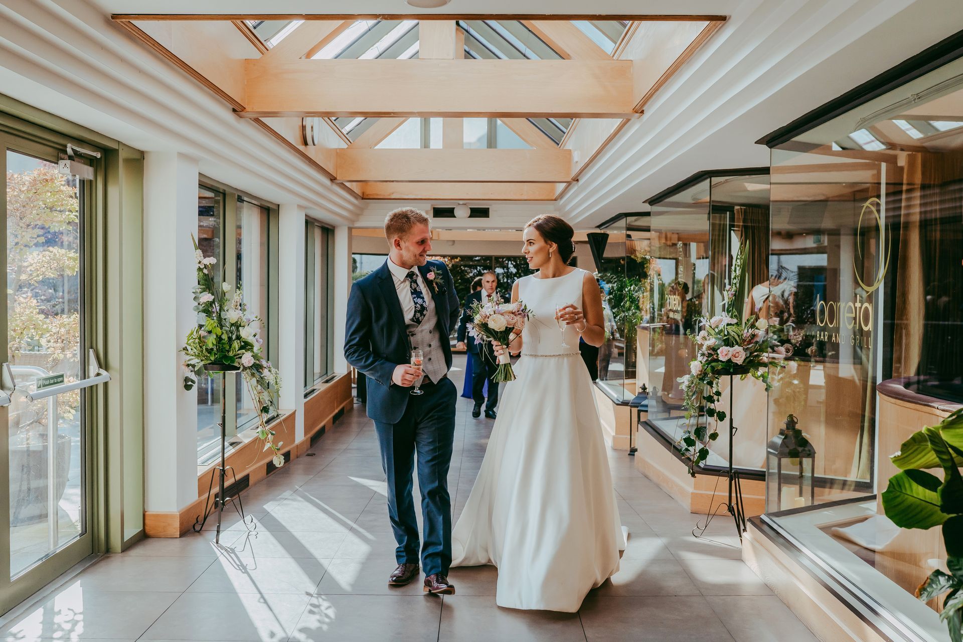 Bride and groom walking together in a bright glass hallway with flowers and skylights