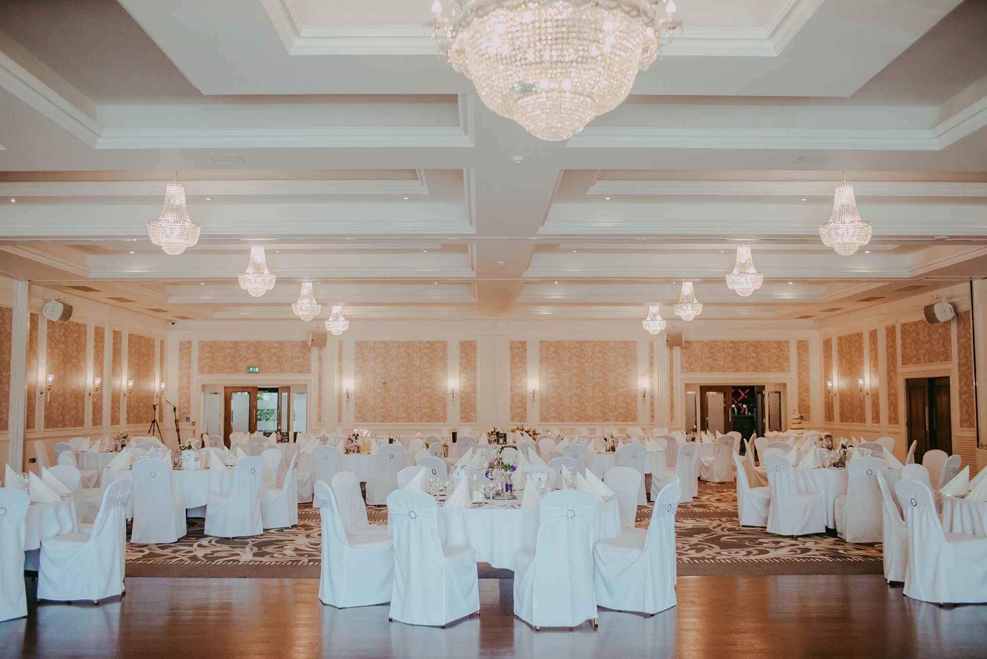 Elegant banquet hall with white-covered chairs, round tables, chandeliers, and warm gold decor