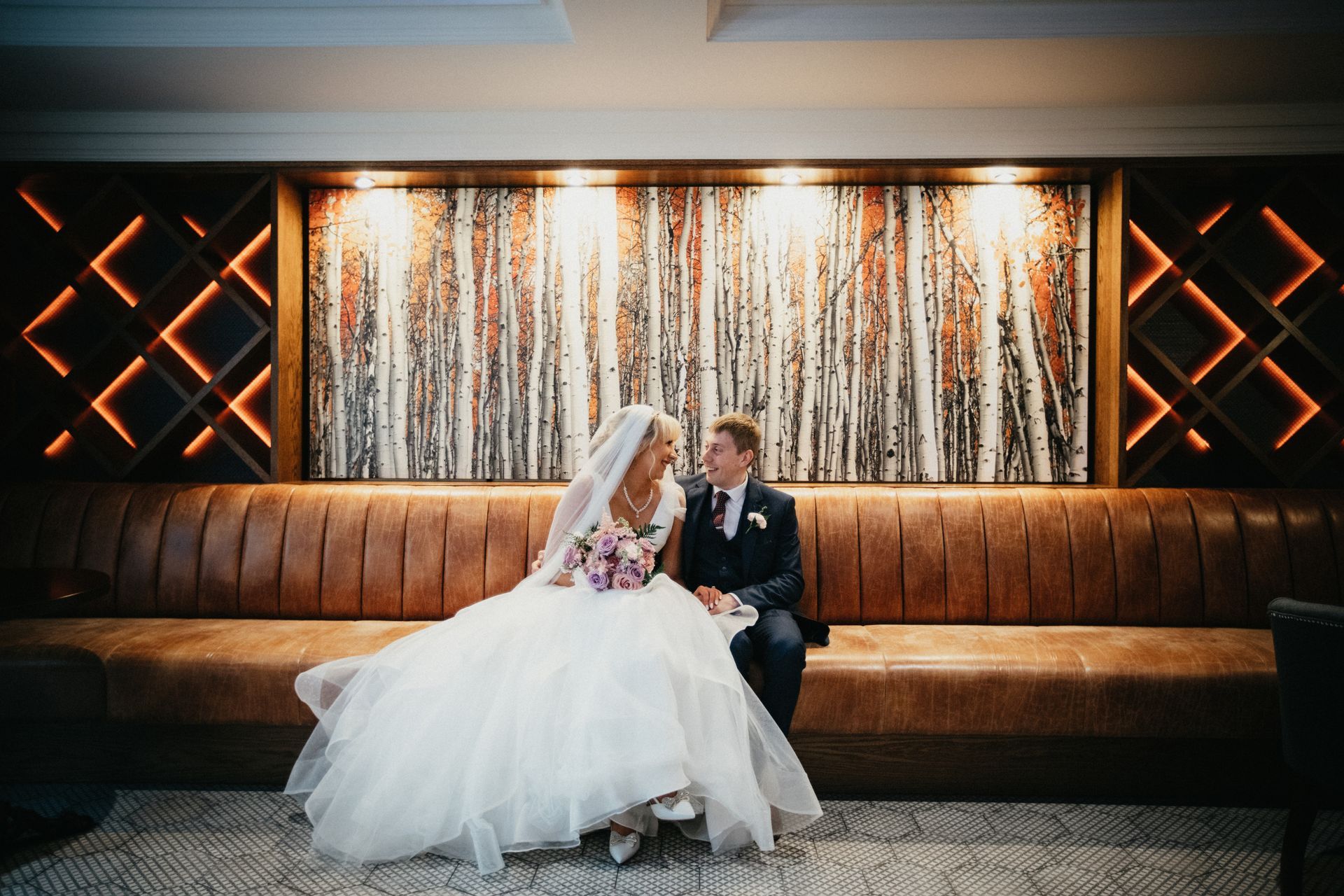 Bride and groom seated on a couch in a warm-lit wedding venue, kissing beside a shimmering backdrop