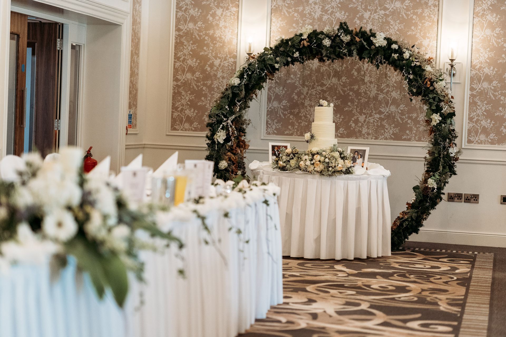 Elegant wedding reception setup with white table linens and a floral arch in a carpeted banquet hall