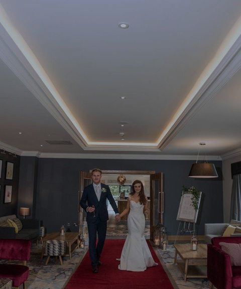 Bride and groom walking hand in hand in a dimly lit elegant room with framed art and red chairs