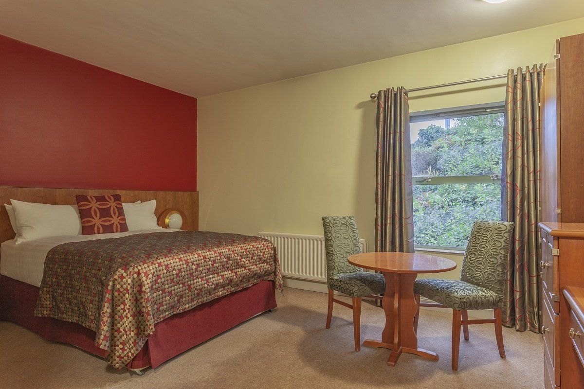 Cozy bedroom with a red accent wall, queen bed, and small round table by a window with chairs.