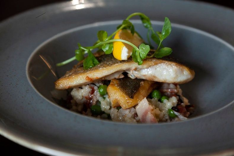 Plated fish fillet on a bed of grains with greens and a lemon garnish in a dark bowl