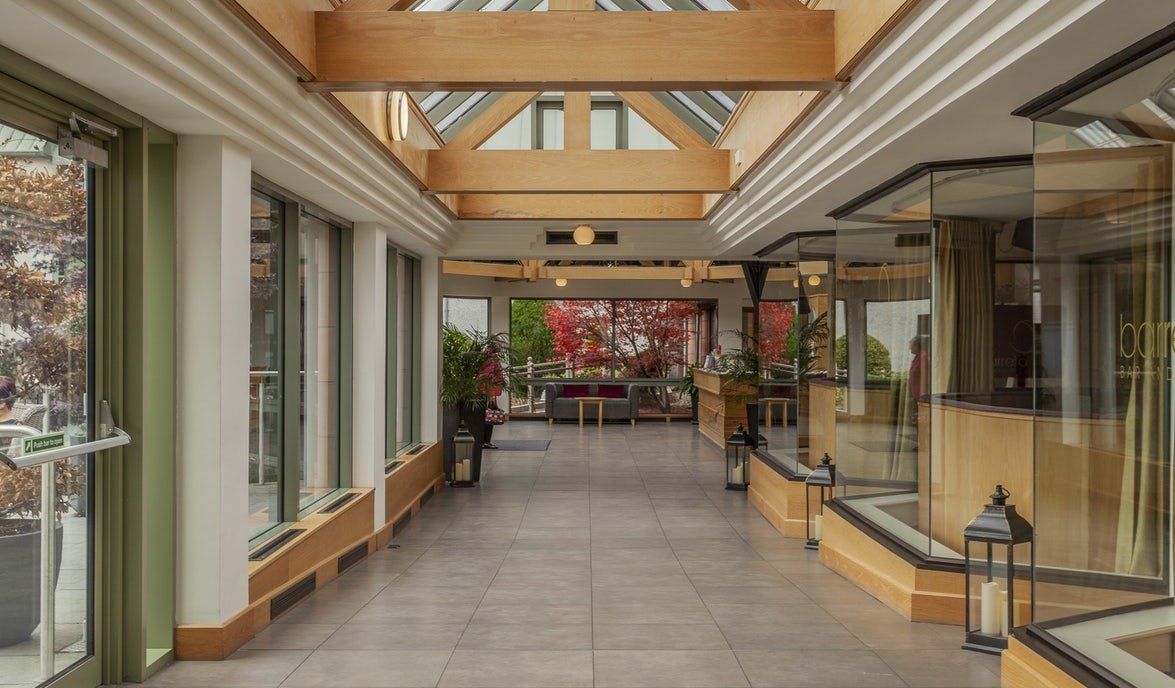 Glass-roofed corridor with large windows, potted plants, and storefronts along both sides