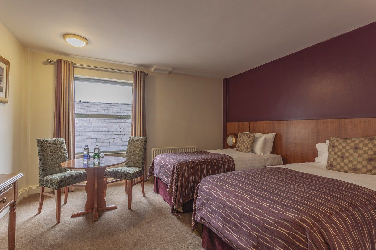Hotel room with two beds, a small table and chairs by a window, and purple accent wall