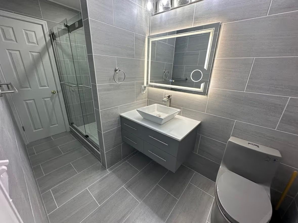 Bathroom remodel in Finksburg with full wall tile, walk-in shower with sliding door, floating vanity, bowl sink, lighted mirror, and gray one-piece toilet