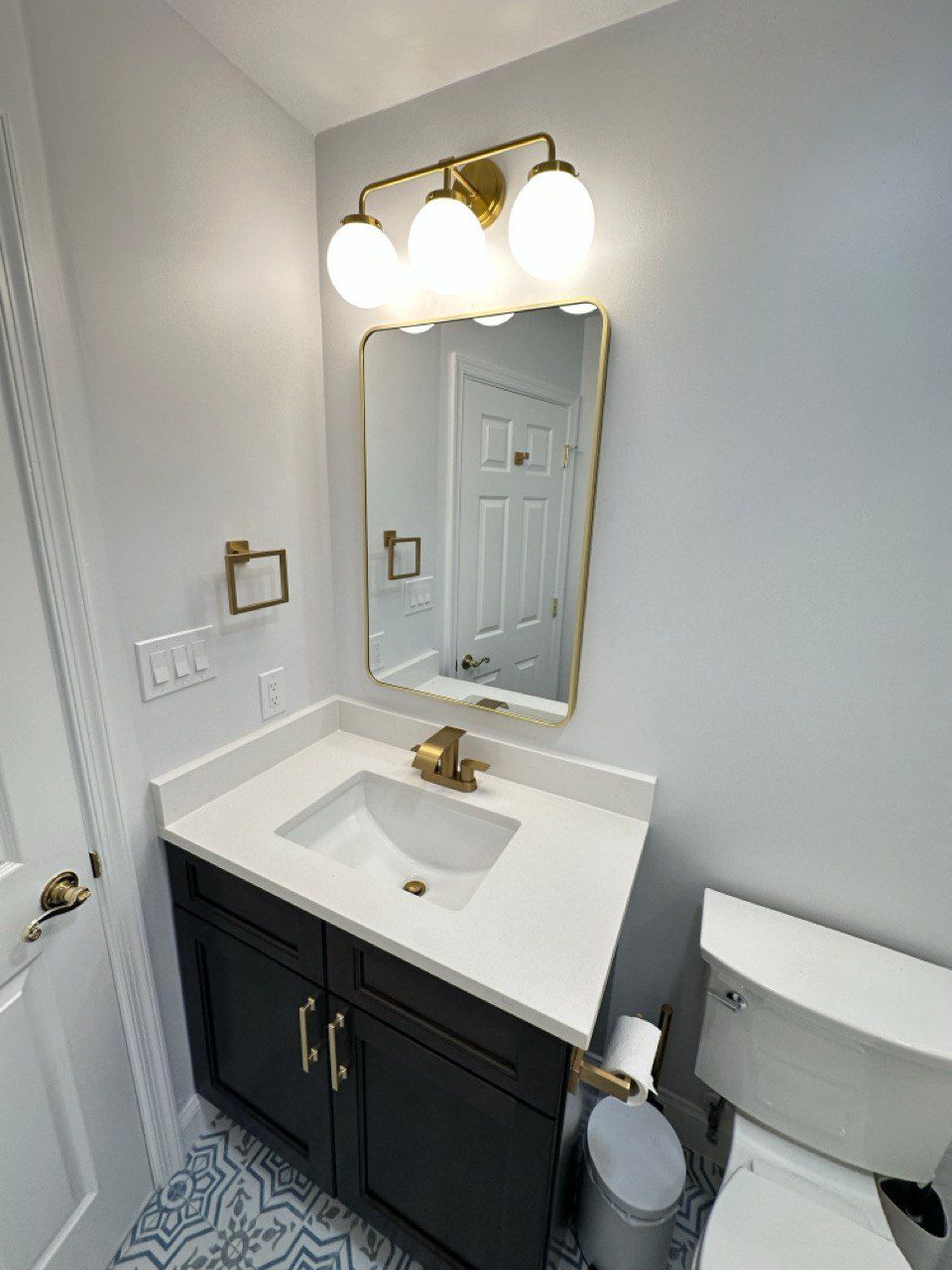 Hallway bathroom remodeling in Sykesville featuring white wavy wall tile with a large horizontal niche accented in blue tile, deep soaking tub with brushed gold faucet and handheld, rain shower head, fixed glass panel, blue floor tile design, custom vanity with quartz top, and a quiet exhaust fan.
