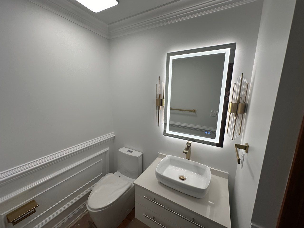 Powder room with floating vanity, bowl sink, brushed gold fixtures, LED mirror, sconces, one-piece toilet, vane scotting, and hardwood flooring