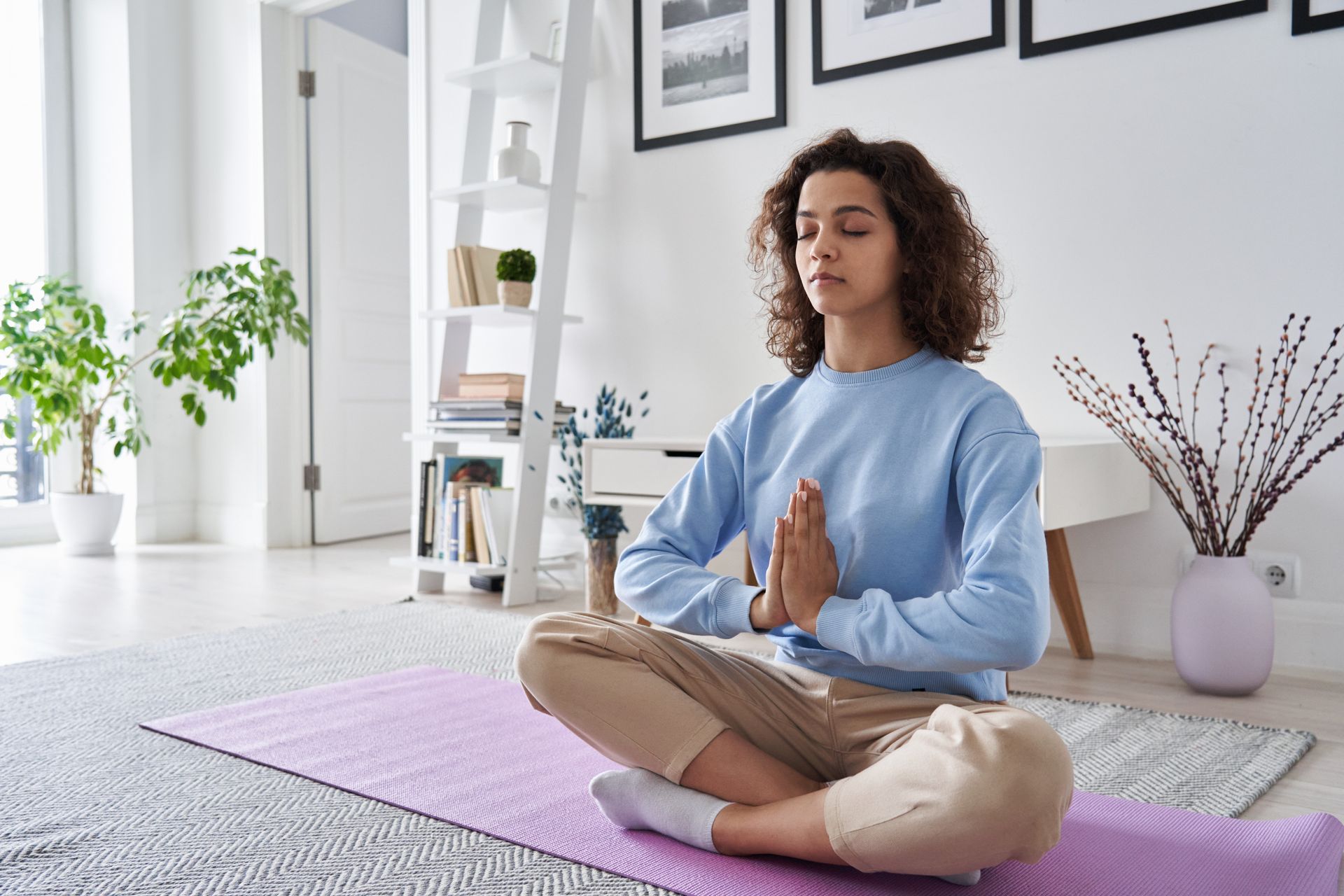 Woman meditating indoors with hands together; sitting on a purple mat. Woman meditating indoors with hands together; sitting on a purple mat.