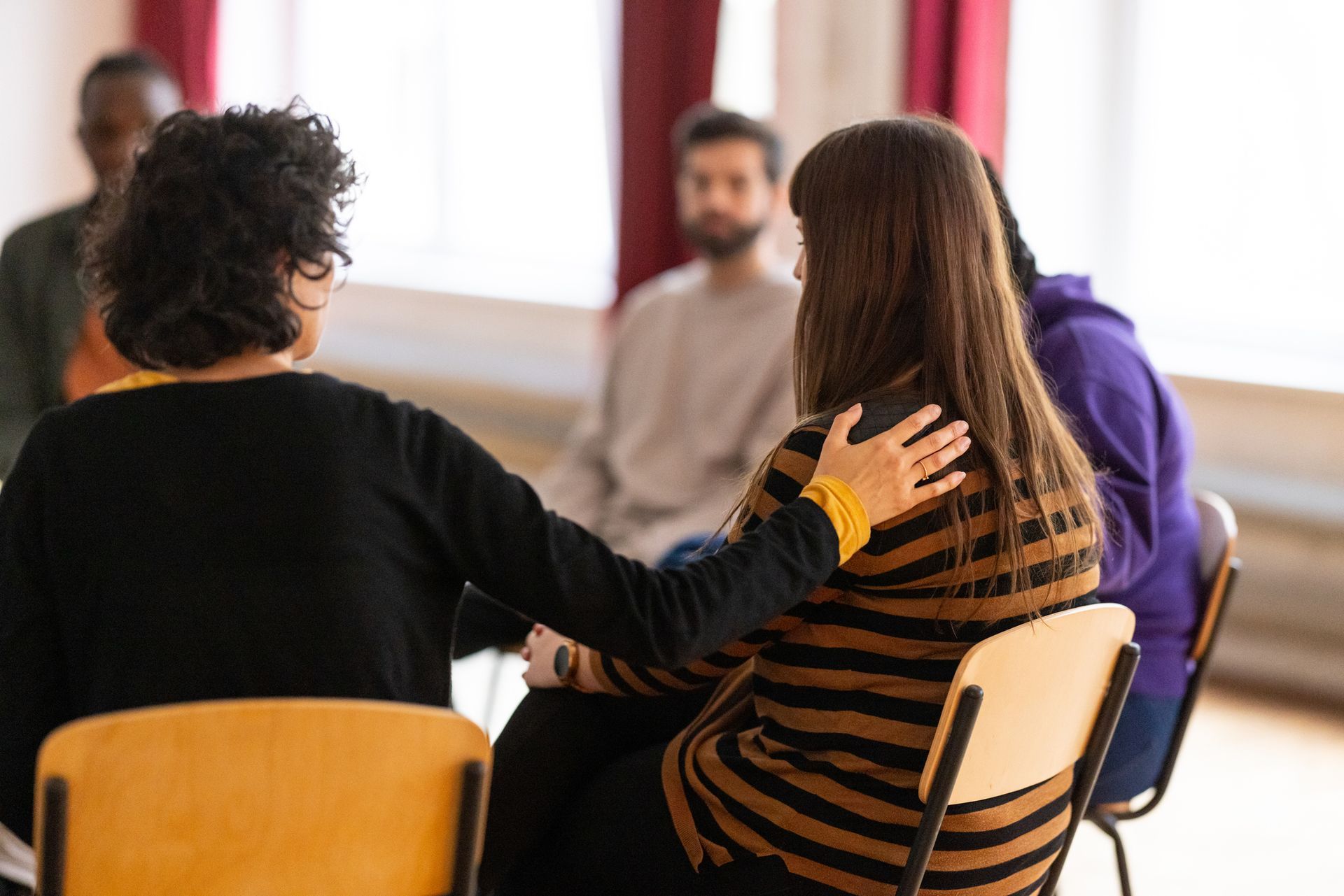 Woman comforts another in a support group, hand on shoulder; other people in a circle.