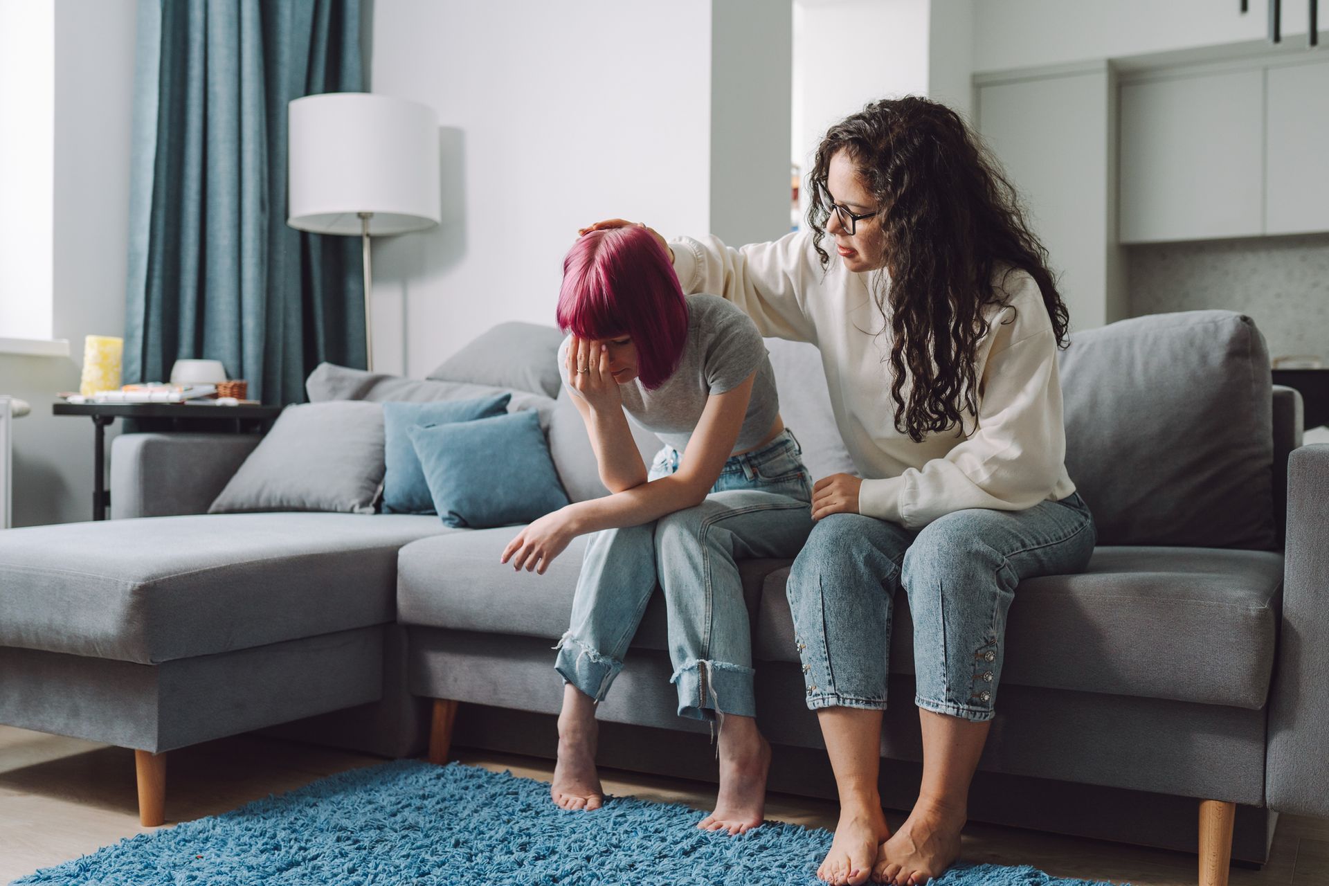 Woman comforts a person, both sitting on a couch. The other person is visibly upset with their head in their hands.