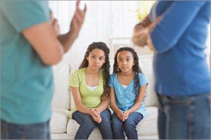 Two girls watch their parents argue. The girls are on a sofa; parents are blurred.
